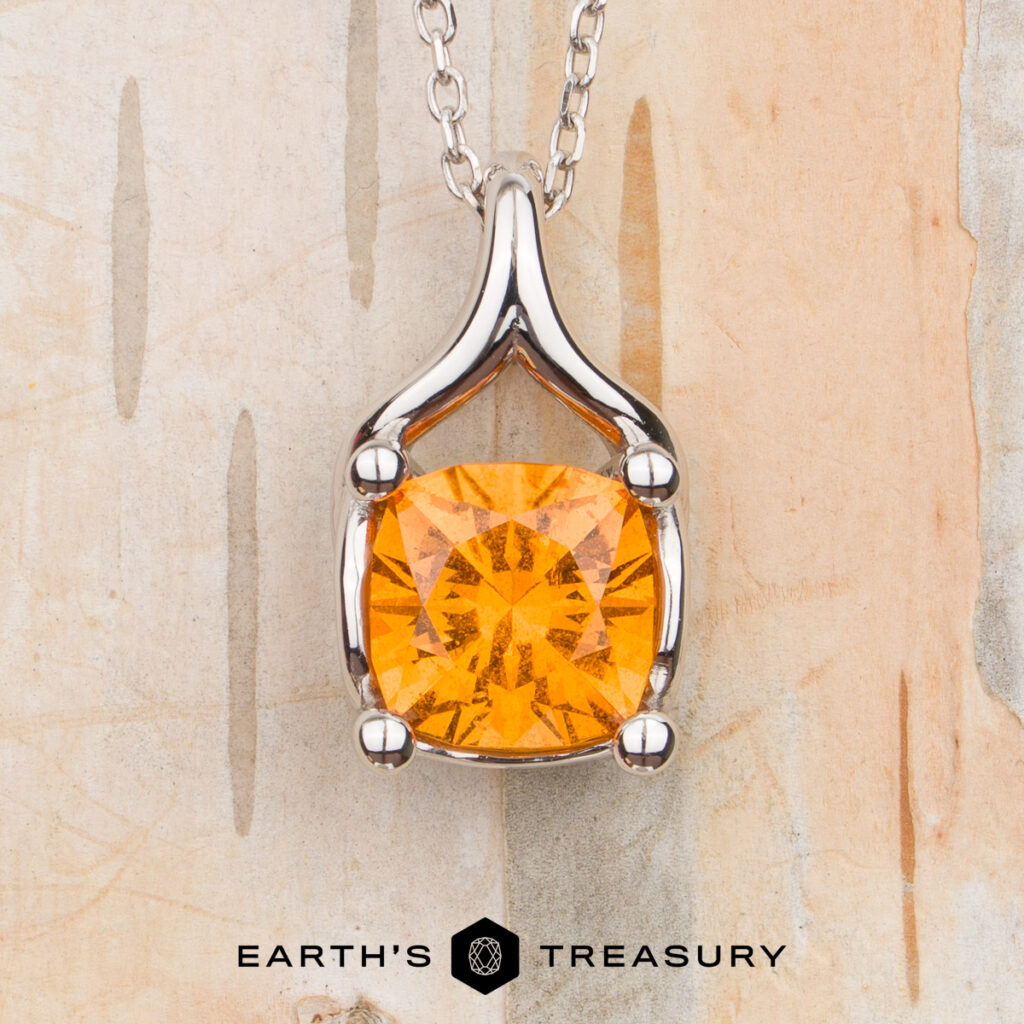 A silver pendant necklace from the Vigh, Melissa 60 [custom gallery] collection, featuring a cushion-cut orange gemstone, is displayed against a light wooden background with the "EARTH'S TREASURY" logo at the bottom.