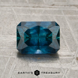 A rectangular, faceted 1.67-carat Peacock Blue Montana Sapphire (Heated) rests on a textured gray surface with the "EARTH'S TREASURY" text and logo displayed below it.