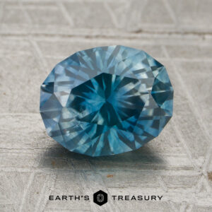 A 2.55-carat faceted oval teal blue Montana sapphire (heated) is placed on a textured gray surface. The logo "Earth's Treasury" appears at the bottom of the image.