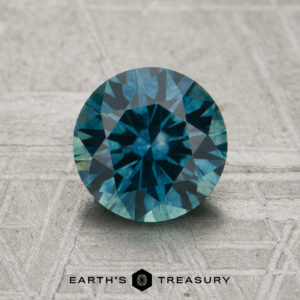 A 1.78-carat teal Montana sapphire (heated) in a round brilliant cut is displayed on a textured surface, with the "EARTH'S TREASURY" logo and text below.