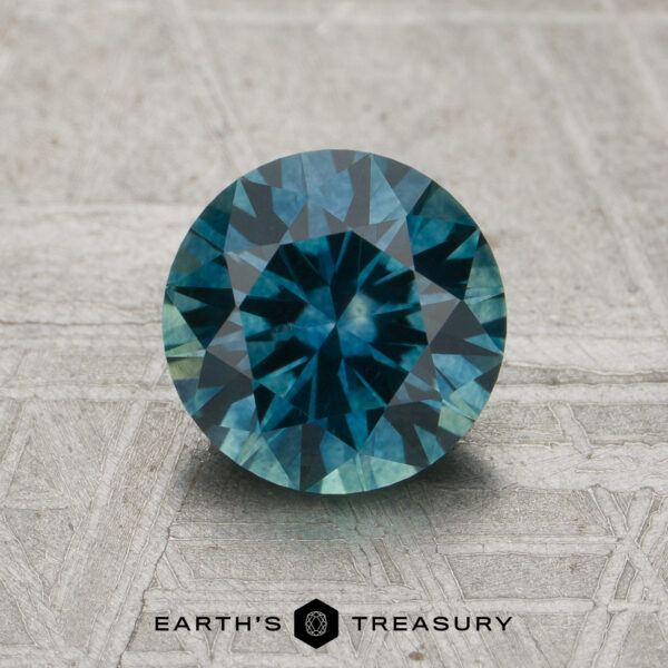 1.38-Carat Teal Montana Sapphire (Heated) A 1.78-carat teal Montana sapphire (heated) in a round brilliant cut is displayed on a textured surface, with the "EARTH'S TREASURY" logo and text below.