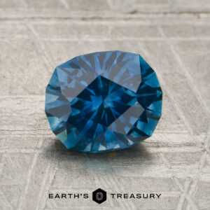 A 1.74-carat rich teal blue Montana sapphire (heated) rests on a textured surface, with the words "Earth's Treasury" and a logo visible at the bottom of the image.