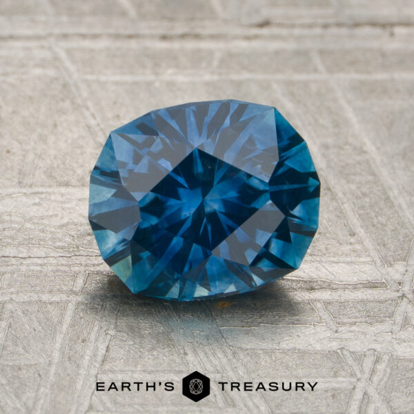A 1.74-carat rich teal blue Montana sapphire (heated) rests on a textured surface, with the words "Earth's Treasury" and a logo visible at the bottom of the image.