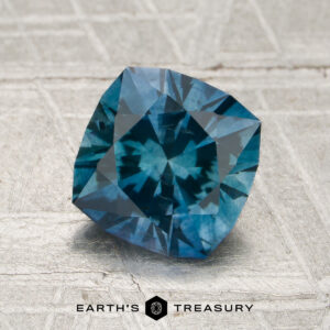 A 1.75-carat heated Montana sapphire, cushion-cut and blue in color, is displayed on a textured gray surface with the Earth's Treasury logo and text visible at the bottom.