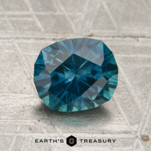 A 1.74-carat teal Montana sapphire (heated) rests on a textured metallic surface featuring the "EARTH'S TREASURY" name and hexagon logo below.