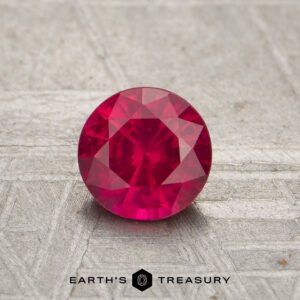 A 0.67-carat red Mozambique ruby (heated), round and faceted, rests on a textured surface with "EARTH'S TREASURY" shown below.