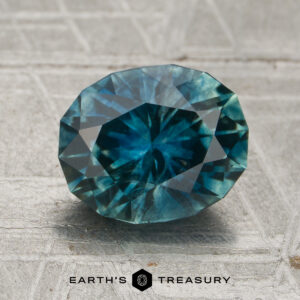 A 2.45-carat teal Montana sapphire (heated), expertly faceted, rests on a textured surface with the Earth's Treasury logo visible at the bottom of the image.