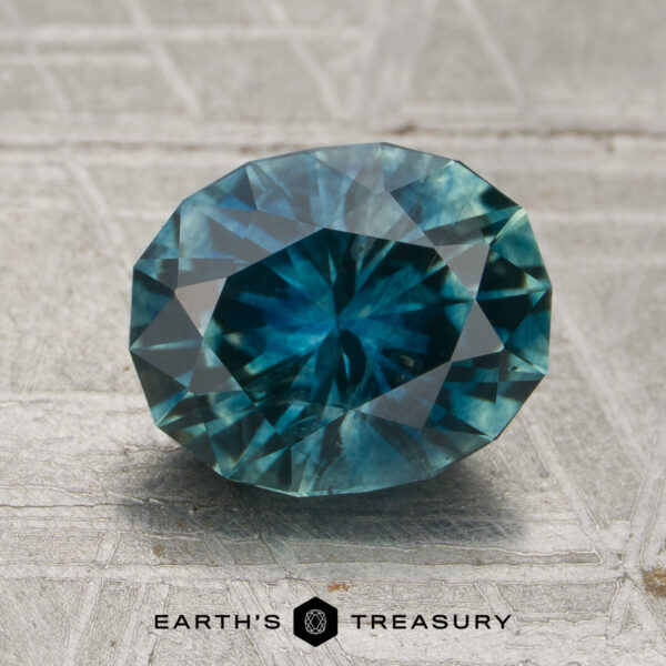 2.45-Carat Teal Montana Sapphire (Heated) A 2.45-carat teal Montana sapphire (heated), expertly faceted, rests on a textured surface with the Earth's Treasury logo visible at the bottom of the image.