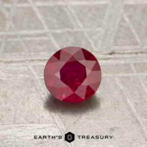 0.56-carat Mozambique Ruby (heated), round-cut, displayed on a textured gray surface with the "Earth's Treasury" logo and text at the bottom of the image.