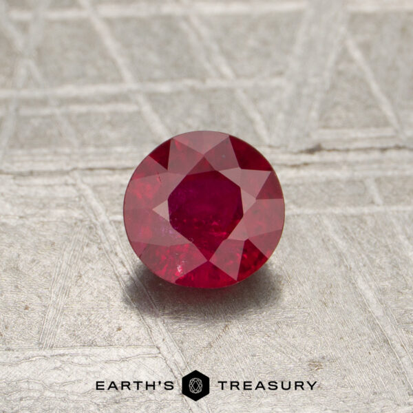 0.56-Carat Mozambique Ruby (Heated) 0.56-carat Mozambique Ruby (heated), round-cut, displayed on a textured gray surface with the "Earth's Treasury" logo and text at the bottom of the image.