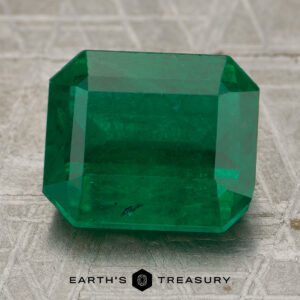 A 5.01-carat rich green Zambian emerald with step-cut facets is displayed on a textured surface, with the Earth's Treasury logo beneath it.
