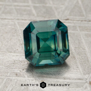 A 1.58-carat square-cut teal sapphire with emerald-style facets rests on textured silver. "EARTH'S TREASURY" is displayed at the bottom.