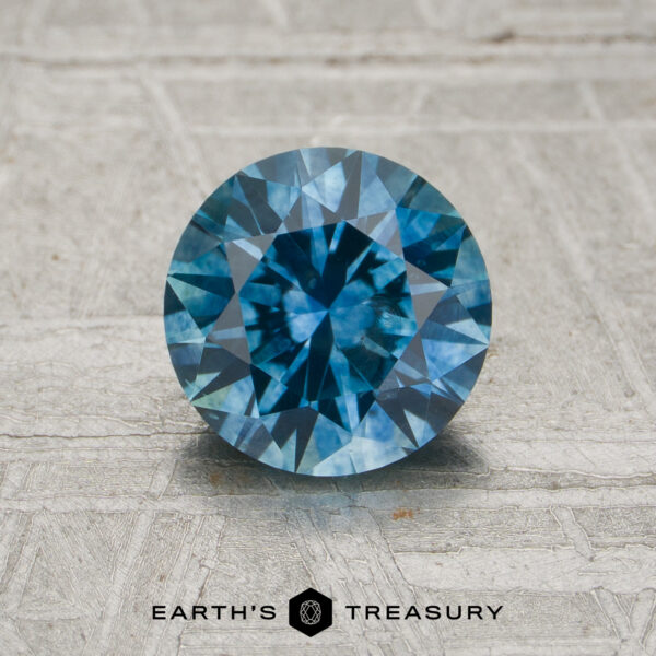 A 1.25-carat medium blue Montana sapphire (heated), round brilliant-cut, is displayed on a textured surface with the text "EARTH'S TREASURY" beneath it.