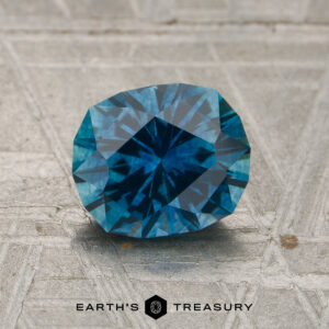A 1.65-carat rich teal blue Montana sapphire (heated) rests on a textured gray surface, with the words "EARTH'S TREASURY" and a logo visible at the bottom.