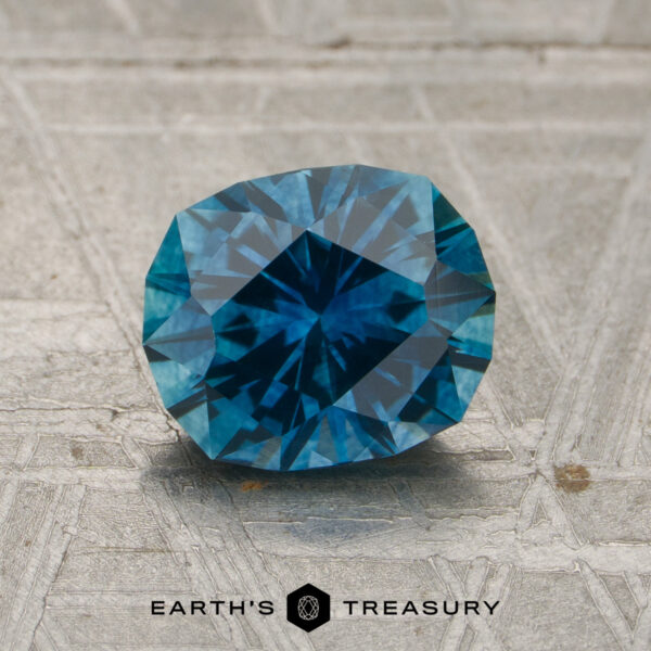 1.65-Carat Rich Blue Montana Sapphire (Heated) A 1.65-carat rich teal blue Montana sapphire (heated) rests on a textured gray surface, with the words "EARTH'S TREASURY" and a logo visible at the bottom.