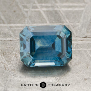 A 2.04-carat teal blue Montana sapphire (heated), featuring a rectangular step cut, is displayed on a textured metallic surface with the “Earth’s Treasury” logo and text at the bottom.