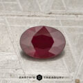 A 0.79-carat heated Burmese ruby, cut in a faceted oval shape, is displayed on a textured gray surface with the Earth's Treasury logo and text at the bottom of the image.