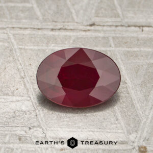 A 0.79-carat heated Burmese ruby, cut in a faceted oval shape, is displayed on a textured gray surface with the Earth's Treasury logo and text at the bottom of the image.