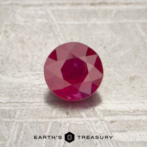 A 0.52-carat red Mozambique ruby (heated), round-cut, displayed on a textured metallic surface with "Earth's Treasury" and a hexagonal logo at the bottom.