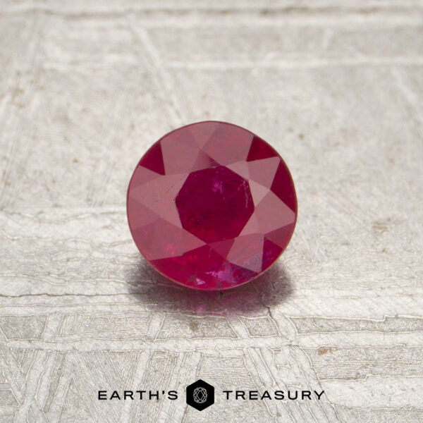 0.52-Carat Red Mozambique Ruby (Heated) A 0.52-carat red Mozambique ruby (heated), round-cut, displayed on a textured metallic surface with "Earth's Treasury" and a hexagonal logo at the bottom.