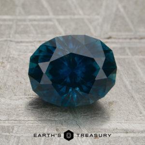 A faceted round 2.31-carat deep peacock blue Montana sapphire (heated) rests on a textured surface. The logo and text "EARTH'S TREASURY" appear at the bottom of the image.