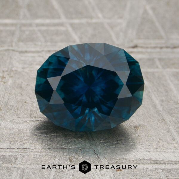 2.31-Carat Deep Peacock Blue Montana Sapphire (Heated) A faceted round 2.31-carat deep peacock blue Montana sapphire (heated) rests on a textured surface. The logo and text "EARTH'S TREASURY" appear at the bottom of the image.