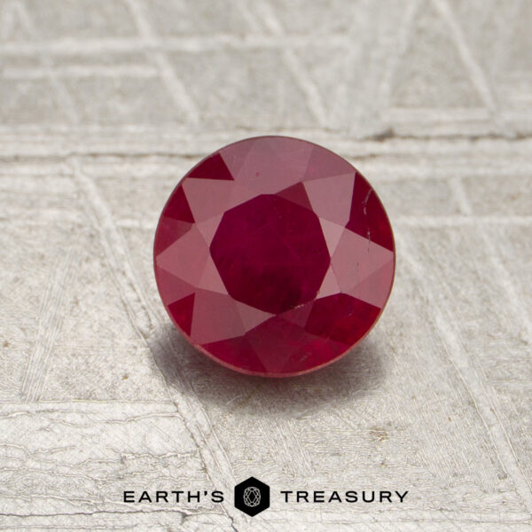 0.77-Carat Mozambique Ruby (Heated) A 0.77-carat heated Mozambique ruby, expertly faceted and round in shape, is displayed on a textured surface with the words "EARTH'S TREASURY" at the bottom.
