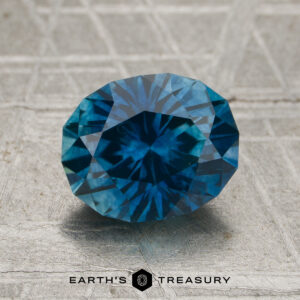 A 2.18-carat rich teal blue Montana sapphire (heated), cut in a faceted oval shape, is displayed on a textured gray surface with the Earth’s Treasury logo beneath it.