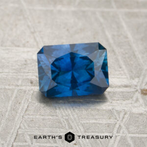 A 1.00-carat rich blue Montana sapphire (heated), cut in a rectangular faceted shape, rests on a textured gray surface. The logo and text "EARTH'S TREASURY" are displayed at the bottom center.