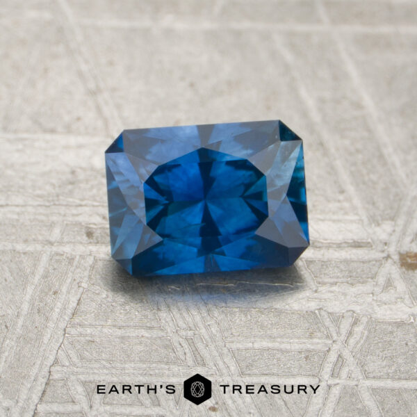 1.00-Carat Rich Blue Montana Sapphire (Heated) A 1.00-carat rich blue Montana sapphire (heated), cut in a rectangular faceted shape, rests on a textured gray surface. The logo and text "EARTH'S TREASURY" are displayed at the bottom center.