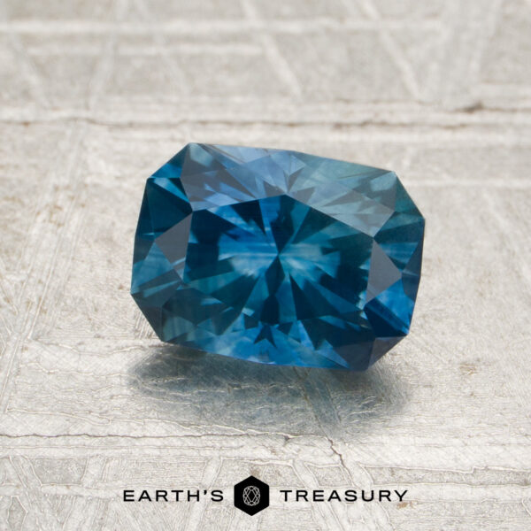 A 1.38-carat heated Montana sapphire, cut in a cushion shape, is displayed on a textured surface with the "Earth's Treasury" logo beneath it.