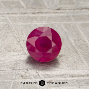 A 0.55-carat red Mozambique ruby (heated), round-cut, is displayed on a textured metallic surface with "EARTH'S TREASURY" and a hexagonal logo below.