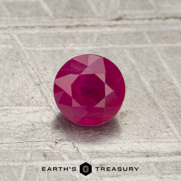 0.55-Carat Red Mozambique Ruby (Heated) A 0.55-carat red Mozambique ruby (heated), round-cut, is displayed on a textured metallic surface with "EARTH'S TREASURY" and a hexagonal logo below.