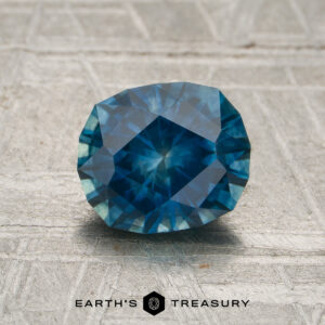 A 1.60-carat rich blue-aqua particolored Montana sapphire, expertly faceted and oval-cut (heated), is displayed on a textured surface. The Earth's Treasury logo and name appear at the bottom.