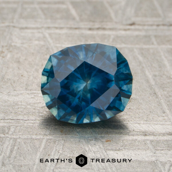 1.60-Carat Rich Blue-Aqua Particolored Montana Sapphire (Heated) A 1.60-carat rich blue-aqua particolored Montana sapphire, expertly faceted and oval-cut (heated), is displayed on a textured surface. The Earth's Treasury logo and name appear at the bottom.