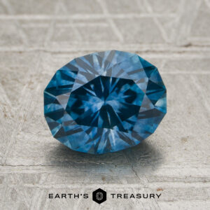 A 2.06-carat medium blue Montana sapphire (heated) rests on a textured gray surface, with the words "Earth's Treasury" and a hexagon logo displayed below it.