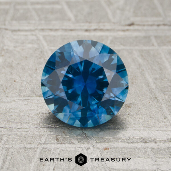 1.21-Carat Rich Blue Montana Sapphire (Heated) shown upright in a round brilliant cut on a textured light-colored surface, with the Earth’s Treasury logo displayed at the bottom.