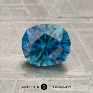 A 1.55-carat Montana rich teal-blue particolored sapphire (heated), cut in an oval faceted shape, is displayed on a gray textured surface, with the "EARTH’S TREASURY" logo and text visible at the bottom.