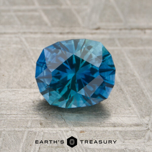 A 1.55-carat Montana rich teal-blue particolored sapphire (heated), cut in an oval faceted shape, is displayed on a gray textured surface, with the "EARTH’S TREASURY" logo and text visible at the bottom.