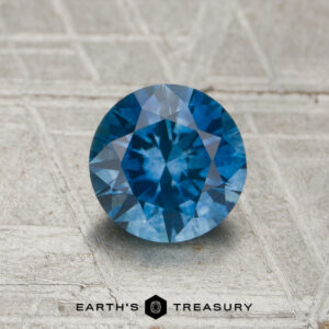 1.10-carat medium blue Montana sapphire (heated), round brilliant-cut, displayed on a textured gray surface with the "Earth's Treasury" logo and text at the bottom.