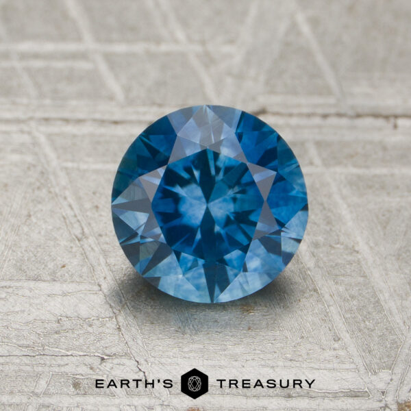 1.10-carat medium blue Montana sapphire (heated), round brilliant-cut, displayed on a textured gray surface with the "Earth's Treasury" logo and text at the bottom.