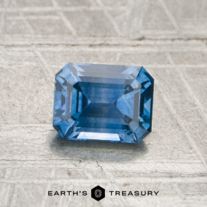 A 1.18-carat medium blue Montana sapphire, featuring an emerald cut, rests on a textured gray surface. The text "EARTH'S TREASURY" appears at the bottom of the image.