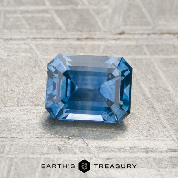 1.18-Carat Medium Blue Montana Sapphire (Heated) A 1.18-carat medium blue Montana sapphire, featuring an emerald cut, rests on a textured gray surface. The text "EARTH'S TREASURY" appears at the bottom of the image.