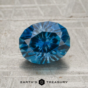 A 2.03-carat rich blue Montana sapphire (heated), faceted in an oval shape, is displayed on a textured light gray surface, with the "Earth's Treasury" logo and text at the bottom.