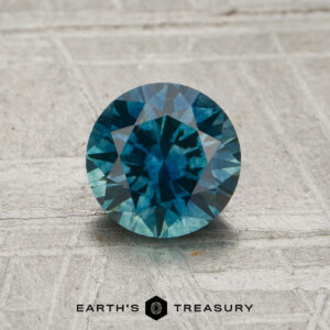 A round, faceted 1.08-carat peacock blue-teal particolored Montana sapphire (heat) rests on a textured gray surface. The Earth’s Treasury logo and text are displayed at the bottom of the image.