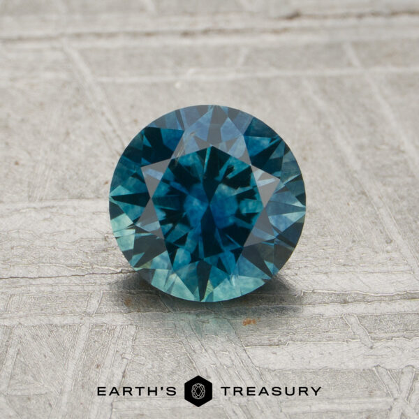 1.08-Carat Peacock Blue-Teal Particolored Montana Sapphire (Heated) A round, faceted 1.08-carat peacock blue-teal particolored Montana sapphire (heat) rests on a textured gray surface. The Earth’s Treasury logo and text are displayed at the bottom of the image.