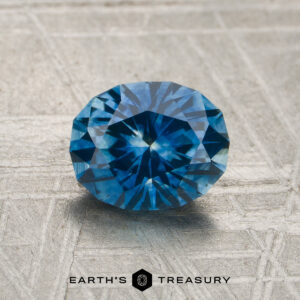 A 1.49-carat rich blue Montana sapphire (heated) is displayed on a textured gray surface, with the logo and text "Earth's Treasury" at the bottom of the image.