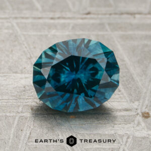 A 2.02-carat rich teal Montana sapphire (heated) is placed on a textured gray surface with the text "EARTH'S TREASURY" and a logo visible below the stone.