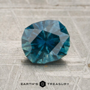 A 1.47-carat heated teal Montana sapphire, faceted in an oval shape, rests on a textured surface with the Earth's Treasury logo and text visible below.