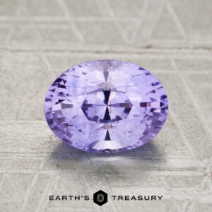 A 1.81-carat faceted oval Ceylon sapphire with a lavender hue rests on a textured light gray surface, with the "Earth’s Treasury" logo displayed at the bottom.